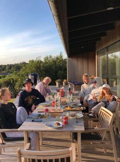 a group of people sitting around a wooden table on top of a deck eating food