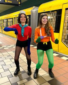 two women dressed in costumes standing next to each other on a subway platform with a yellow train behind them