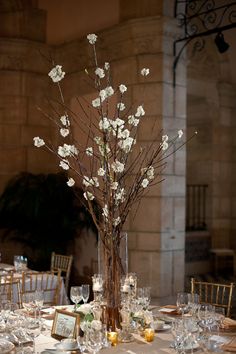 a tall vase with white flowers on top of a table filled with glasses and place settings