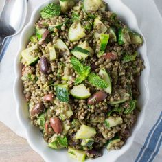 a white bowl filled with lentils, cucumbers and broccoli on top of a wooden table