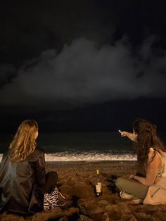 two women sitting on the beach pointing at something in the sky with dark clouds behind them