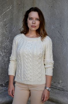 a woman standing in front of a stone wall wearing a white sweater and khaki pants