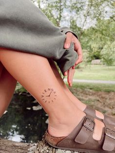 a woman's foot with a small compass tattoo on her left side leg, sitting next to a body of water