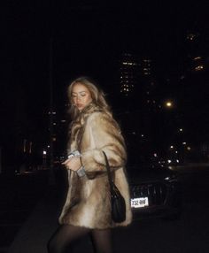 a woman in a fur coat is walking down the street at night with her cell phone