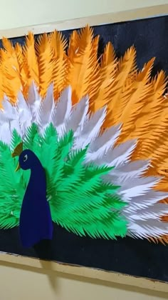 a peacock made out of paper sitting on top of a black board with orange, white and green feathers