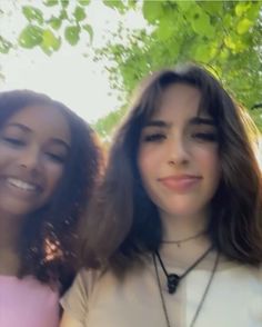 two women standing next to each other in front of some trees and green leaves on a sunny day