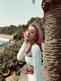 a woman with pink hair standing next to a palm tree near the ocean and beach