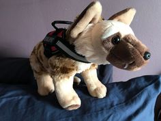 a stuffed dog with a harness on sitting on a blue bed sheet in front of a purple wall