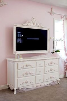 a white dresser with a tv on top of it next to a window in a pink room