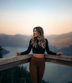 a woman standing on top of a wooden balcony next to a body of water with mountains in the background