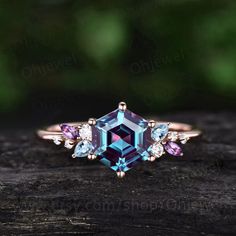 an engagement ring with blue and pink stones on top of a piece of wood in front of green leaves