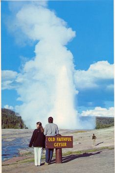 two people standing in front of a geyser with a blue sky and clouds