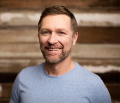 a man with a beard and blue shirt smiles at the camera while standing in front of wooden planks