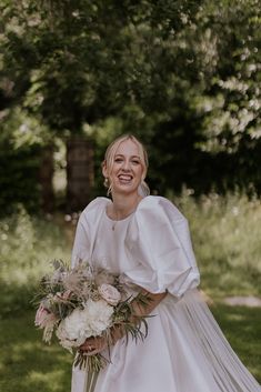 a woman in a white wedding dress holding a bouquet and smiling at the camera with trees behind her