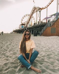 a woman sitting in the sand near a roller coaster at an amusement park or beach