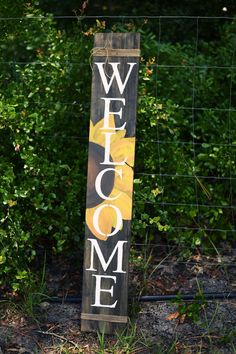 a welcome sign in front of a fence with bushes behind it and the word welcome painted on it