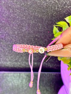 a hand holding a pink beaded bracelet with gold beads on it and a flower pot in the background