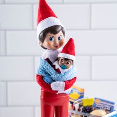 an elf holding a baby in his arms on top of a red plastic container filled with candy