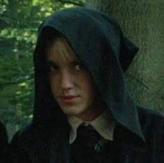 two young men standing next to each other in the woods, one wearing a hoodie