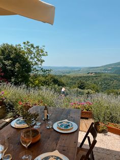 an outdoor table with plates and wine glasses on it in front of a scenic view