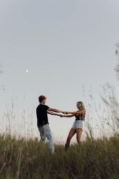 two people standing in tall grass holding hands and looking at the sky with a half moon behind them