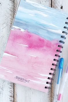 a pink and blue watercolored notebook next to a pen on a wooden table