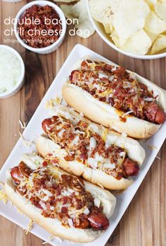 three hot dogs with toppings sit on a plate next to bowls of potato chips