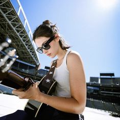 a woman sitting on the ground holding a guitar and looking at her cell phone while wearing sunglasses