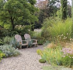 two wooden chairs sitting in the middle of a graveled area surrounded by plants and flowers