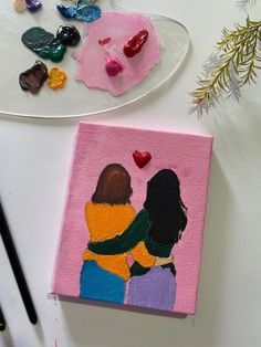 two women hugging each other on a pink canvas next to some paint and pencils