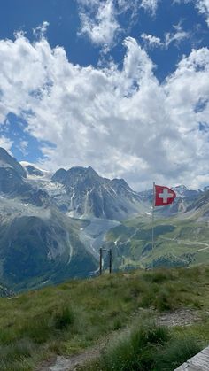 a flag on top of a grassy hill with mountains in the background and clouds in the sky