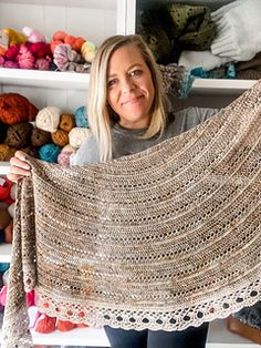 a woman holding up a crocheted shawl in front of shelves full of yarn