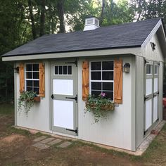 a small shed with two windows and shutters on the doors is shown in front of some trees