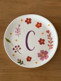 a plate with the letter c painted on it and flowers in the center, sitting on a wooden surface