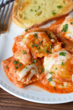 a white plate topped with meat and cheese covered ravioli next to a slice of bread