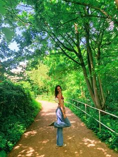 a woman standing in the middle of a dirt road with trees around her and one hand on her hip