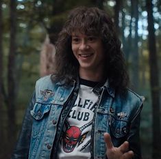 a man with long hair wearing a jean jacket and holding his hand up in the air