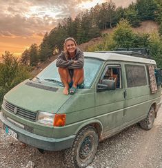 a woman sitting on top of a green van in the middle of a dirt road