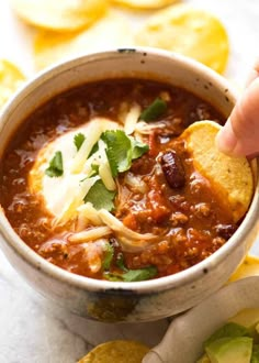 a hand dipping tortilla chips into a bowl of chili