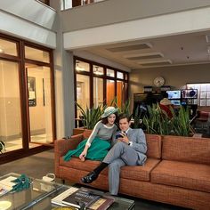 a man and woman are sitting on a couch in an office building with plants behind them