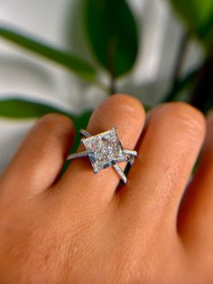 a woman's hand with a diamond ring on top of her finger, next to a potted plant