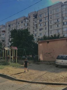 a little boy walking across a dirt field next to a white car and tall buildings