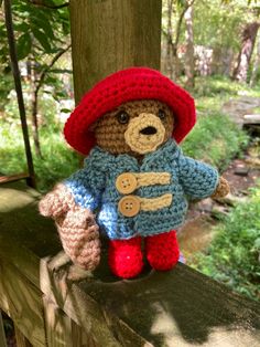 a crocheted teddy bear wearing a red hat and blue jacket sitting on a fence