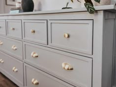 a white dresser with gold knobs in a living room or dining room, next to a plant