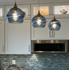 three lights hanging from the ceiling above a sink in a kitchen with white cabinets and stainless steel faucet