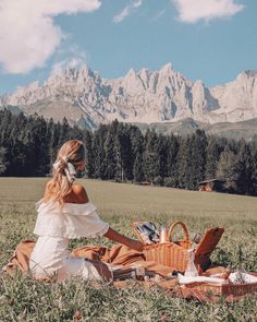 a woman sitting in the grass with a picnic basket on her lap and mountains in the background