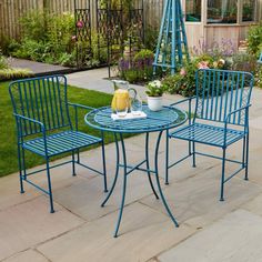 two blue chairs and a table in a backyard with flowers on the side walk way