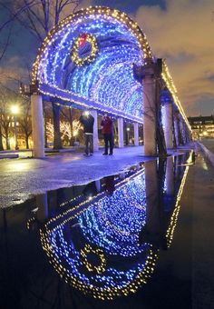 people are standing on the sidewalk with lights all around them and reflecting in the water