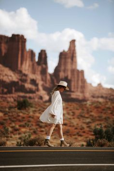 a woman in a white dress and hat walking on the side of a road with mountains in the background