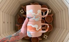 a hand holding a pink coffee mug in front of a stack of brown cups on top of each other